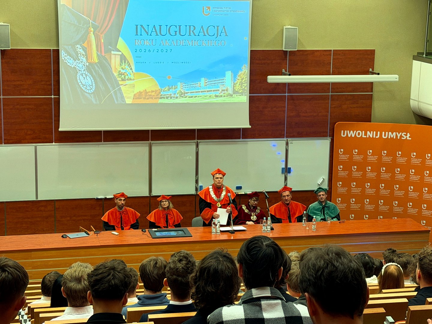 Widok auli podczas uroczystej inauguracji roku akademickiego 2026/2027 na Wydziale Fizyki i Informatyki Stosowanej UŁ. Przy stole prezydialnym siedzą przedstawiciele władz uczelni w togach i biretach, a na środku przemawia Dziekan Wydziału. Na ekranie wyświetla się grafika inauguracji, a na pierwszym planie widać zgromadzonych studentów i gości.