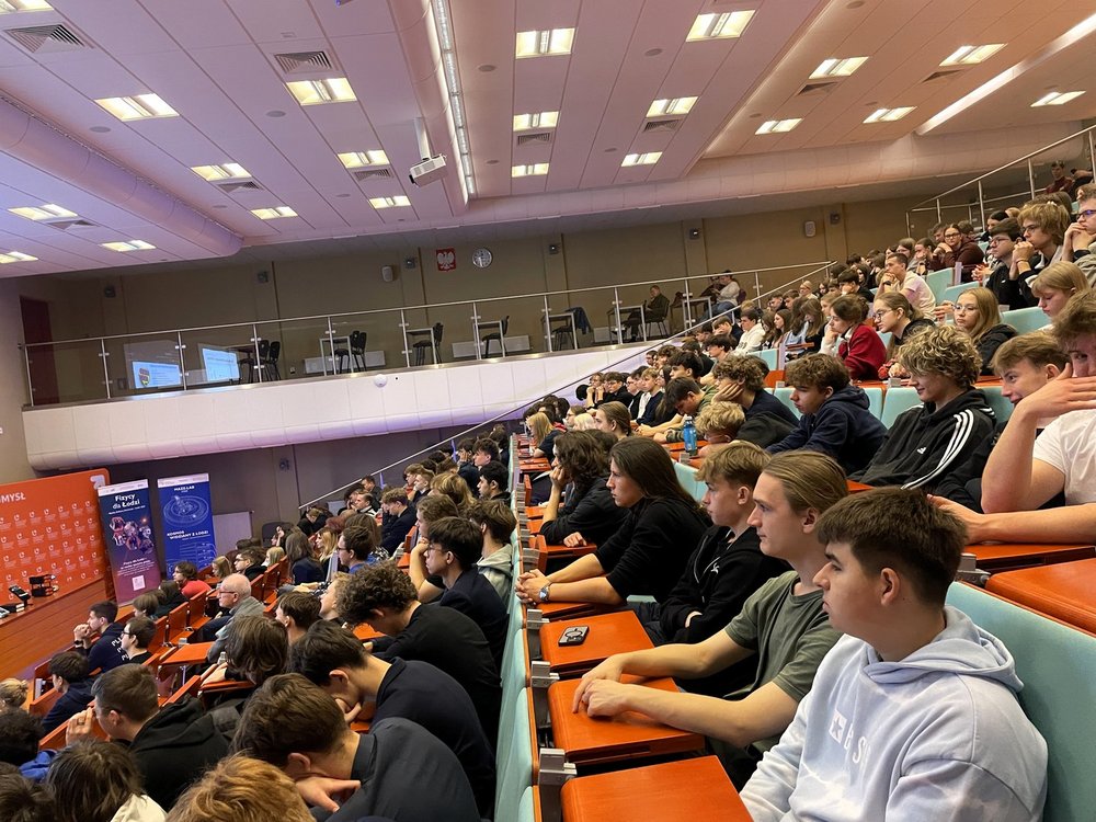 Duża sala wykładowa wypełniona uczniami i uczestnikami wydarzenia. Wszyscy siedzą w rzędach skierowanych w stronę sceny, gdzie prowadzący prezentuje materiał wyświetlany na dwóch dużych ekranach. Widać jasne oświetlenie, drewniane ławki i szeroką przestrzeń audytorium. Uczestnicy uważnie śledzą przebieg wykładu.