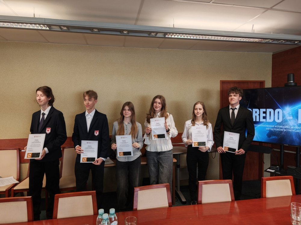 W sali konferencyjnej stoi w jednym rzędzie sześcioro młodych ludzi. Wszyscy trzymają w dłoniach dyplomy i pozują do pamiątkowego zdjęcia. Dwie osoby mają na sobie jasne koszule, pozostali ubrani są bardziej formalnie – w ciemne marynarki lub garnitury. Uczestnicy stoją na tle jasnej ściany, a po prawej stronie znajduje się duży ekran z grafiką o tematyce kosmicznej i napisem „REDO”. Przed grupą widoczny jest długi drewniany stół konferencyjny z krzesłami oraz kilkoma butelkami wody i szklankami. Atmosfera zdjęcia sugeruje uroczyste wręczenie dyplomów lub nagród podczas wydarzenia edukacyjnego albo naukowego.