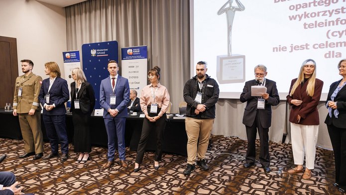 Uczestnicy 32. Forum Teleinformatyki stoją w szeregu podczas uroczystości wręczenia nagród. W tle widoczne są logotypy organizatorów i partnerów wydarzenia oraz ekran prezentacyjny.
