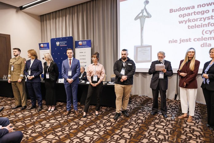 Uczestnicy 32. Forum Teleinformatyki stoją w szeregu podczas uroczystości wręczenia nagród. W tle widoczne są logotypy organizatorów i partnerów wydarzenia oraz ekran prezentacyjny.