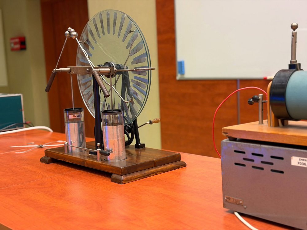 Na stole w sali laboratoryjnej znajduje się zabytkowy elektroskop z dużą okrągłą tarczą i metalowymi elementami, obok którego ustawiono inne urządzenie do doświadczeń z elektryczności. W tle widoczna jest tablica oraz wyposażenie sali dydaktycznej.