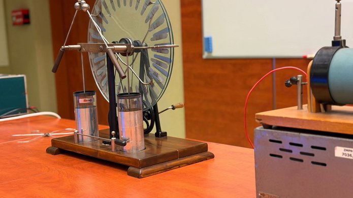 Na stole w sali laboratoryjnej znajduje się zabytkowy elektroskop z dużą okrągłą tarczą i metalowymi elementami, obok którego ustawiono inne urządzenie do doświadczeń z elektryczności. W tle widoczna jest tablica oraz wyposażenie sali dydaktycznej.