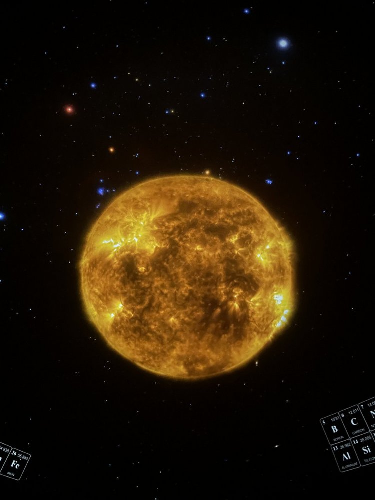 Na środku obrazu znajduje się duża, jasnożółta kula przedstawiająca Słońce. Jego powierzchnia jest nierówna i dynamiczna – widoczne są jaśniejsze i ciemniejsze obszary, przypominające gorące, wijące się struktury plazmy. W kilku miejscach pojawiają się wyraźne, intensywnie świecące punkty, które wyglądają jak rozbłyski słoneczne.  Tło zdjęcia jest czarne i wypełnione licznymi gwiazdami w różnych kolorach: białymi, niebieskimi, pomarańczowymi i czerwonymi. W dolnych rogach kadru widać fragmenty schematycznych tablic – przypominają elementy tablicy okresowej pierwiastków.  Całość przypomina naukową wizualizację lub kadr z filmu edukacyjnego o Słońcu i strukturze Wszechświata.