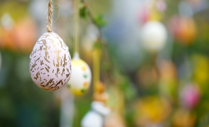 Zbliżenie na wielkanocną dekorację: białą pisankę z nieregularnym złotym wzorem, zawieszoną na sznurku. Jajko znajduje się po lewej stronie obrazu i jest ostre oraz dobrze widoczne.  W tle, które jest rozmyte, wiszą inne kolorowe pisanki zawieszone na gałązkach. Widać pastelowe kolory – żółty, różowy, zielony i biały. Całość tworzy wiosenną, świąteczną atmosferę z dominującymi zielonymi i żółtymi odcieniami tła.