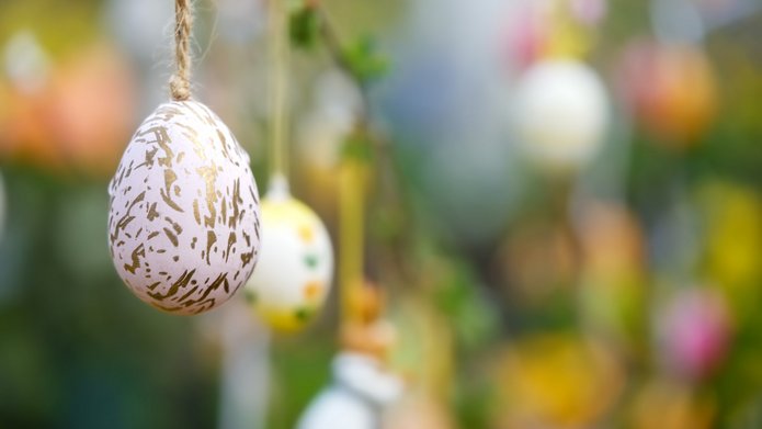 Zbliżenie na wielkanocną dekorację: białą pisankę z nieregularnym złotym wzorem, zawieszoną na sznurku. Jajko znajduje się po lewej stronie obrazu i jest ostre oraz dobrze widoczne.  W tle, które jest rozmyte, wiszą inne kolorowe pisanki zawieszone na gałązkach. Widać pastelowe kolory – żółty, różowy, zielony i biały. Całość tworzy wiosenną, świąteczną atmosferę z dominującymi zielonymi i żółtymi odcieniami tła.