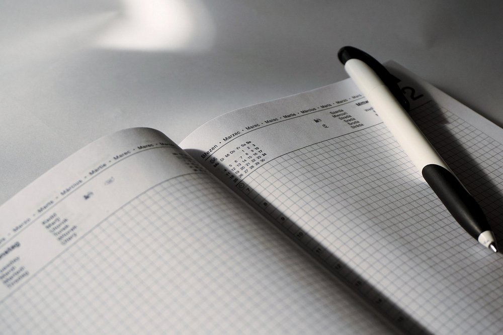 Na zdjęciu widać otwarty notes lub planer leżący na jasnej powierzchni. Obie strony mają delikatną kratkę do pisania. U góry strony widoczny jest nagłówek z nazwami miesięcy w różnych językach, m.in. „Březen”, „Marec”, „März”, „Marzo”, co wskazuje na marzec. Na prawej stronie notesu, ukośnie, leży biało-czarny długopis. Światło pada z lewej strony, tworząc wyraźne cienie długopisu i zagiętej kartki. Całość ma spokojny, minimalistyczny charakter i sugeruje planowanie lub notowanie.