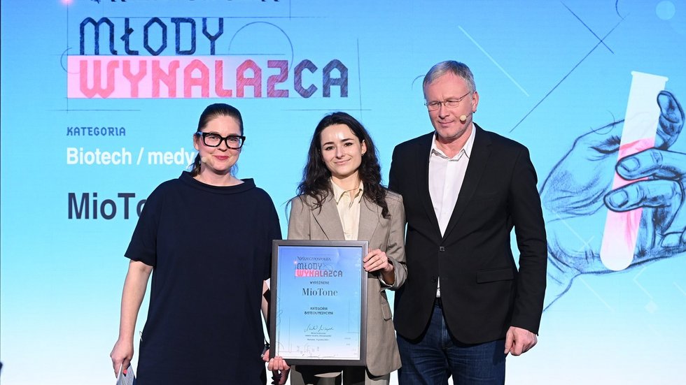 Na zdjęciu widać trzy osoby stojące na scenie podczas uroczystej gali. W centrum znajduje się młoda kobieta ubrana w jasny garnitur, trzymająca w dłoniach oprawiony dyplom. Towarzyszą jej dwie osoby dorosłe — kobieta po lewej stronie i mężczyzna po prawej — ubrane w ciemne, eleganckie stroje. Wszyscy stoją przodem do obiektywu i pozują do zdjęcia.  W tle widoczny jest duży ekran z napisem „Rzeczpospolita – Młody Wynalazca” oraz informacją o kategorii „Biotech / medycyna”. Scena jest profesjonalnie oświetlona, a całość ma oficjalny, galowy charakter.