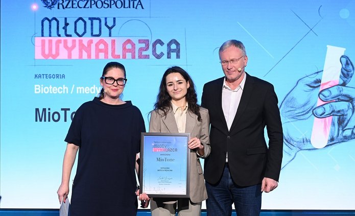 Na zdjęciu widać trzy osoby stojące na scenie podczas uroczystej gali. W centrum znajduje się młoda kobieta ubrana w jasny garnitur, trzymająca w dłoniach oprawiony dyplom. Towarzyszą jej dwie osoby dorosłe — kobieta po lewej stronie i mężczyzna po prawej — ubrane w ciemne, eleganckie stroje. Wszyscy stoją przodem do obiektywu i pozują do zdjęcia.  W tle widoczny jest duży ekran z napisem „Rzeczpospolita – Młody Wynalazca” oraz informacją o kategorii „Biotech / medycyna”. Scena jest profesjonalnie oświetlona, a całość ma oficjalny, galowy charakter.
