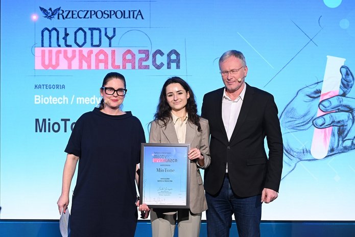 Na zdjęciu widać trzy osoby stojące na scenie podczas uroczystej gali. W centrum znajduje się młoda kobieta ubrana w jasny garnitur, trzymająca w dłoniach oprawiony dyplom. Towarzyszą jej dwie osoby dorosłe — kobieta po lewej stronie i mężczyzna po prawej — ubrane w ciemne, eleganckie stroje. Wszyscy stoją przodem do obiektywu i pozują do zdjęcia.  W tle widoczny jest duży ekran z napisem „Rzeczpospolita – Młody Wynalazca” oraz informacją o kategorii „Biotech / medycyna”. Scena jest profesjonalnie oświetlona, a całość ma oficjalny, galowy charakter.