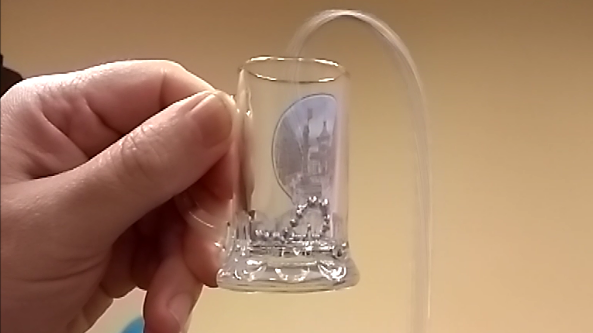 Na zdjęciu widać dłoń trzymającą niewielkie, przezroczyste szklane naczynie przypominające mały kieliszek. Wewnątrz naczynia znajdują się drobne, metaliczne kulki. Z prawej strony do naczynia wprowadzona jest przezroczysta, cienka rurka, która wygina się łukiem nad jego krawędzią i prowadzi na zewnątrz.  Tło zdjęcia jest jednolite i jasne, w kolorze beżowym, dzięki czemu główny element – szklane naczynie z kulkami i rurką – jest dobrze widoczny na pierwszym planie. Całość wygląda jak fragment prostego doświadczenia fizycznego z użyciem małego naczynia, kulek i elastycznej rurki.