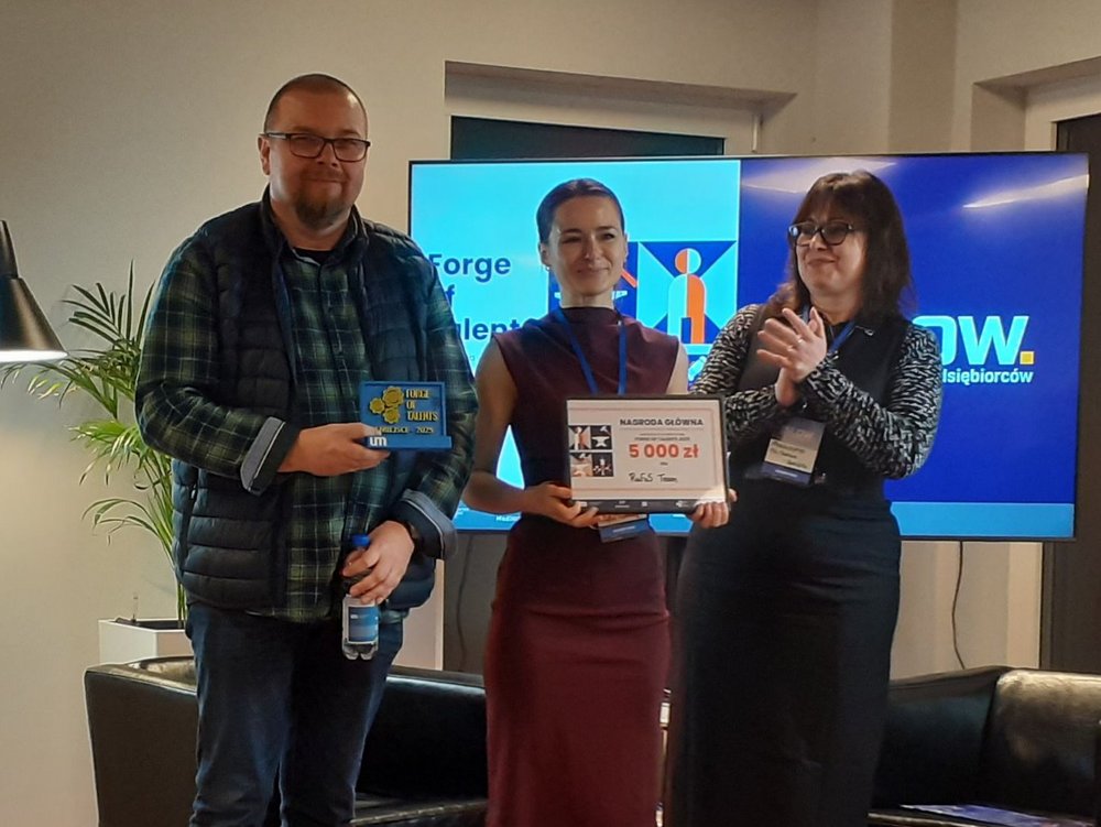 Na zdjęciu troje uczestników konkursu Forge of Talents 2025 stoi na czerwonym dywanie w sali konferencyjnej. Dwoje z nich trzyma dyplom i tabliczkę z napisem „Nagroda Główna 5000 zł”, a obok nich kobieta klaszcze, gratulując zwycięzcom. W tle widać ekran z logo wydarzenia Forge of Talents oraz sprzęt oświetleniowy.