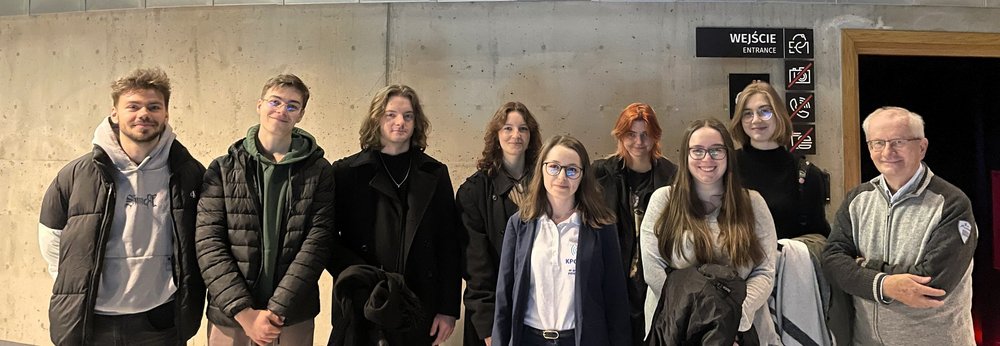 Na zdjęciu widać grupę dziewięciu osób stojących w dwóch rzędach na tle jasnej, betonowej ściany we wnętrzu budynku. Wszyscy patrzą w stronę aparatu i uśmiechają się. Osoby mają na sobie jesienne ubrania – kurtki, swetry i bluzy. W tle, po prawej stronie, znajduje się wejście oznaczone napisem „Wejście / Entrance” oraz kilka piktogramów z informacjami dla odwiedzających. Zdjęcie ma charakter pamiątkowy i przedstawia uczestników wydarzenia stojących wspólnie w grupie.