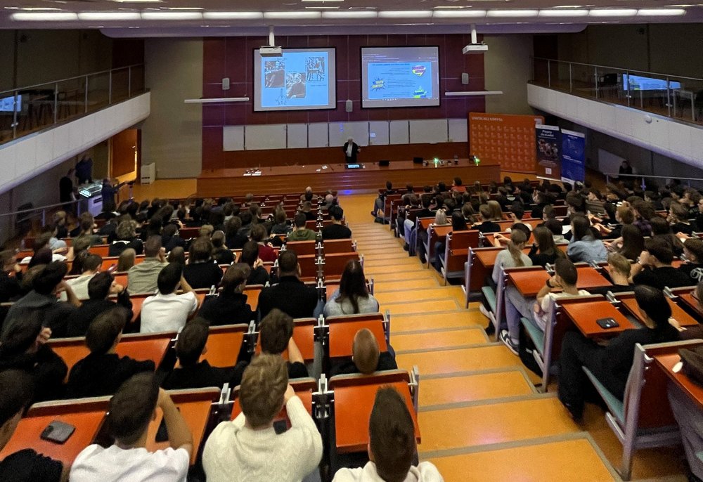 Duża sala wykładowa wypełniona uczniami i uczestnikami wydarzenia. Wszyscy siedzą w rzędach skierowanych w stronę sceny, gdzie prowadzący prezentuje materiał wyświetlany na dwóch dużych ekranach. Widać jasne oświetlenie, drewniane ławki i szeroką przestrzeń audytorium. Uczestnicy uważnie śledzą przebieg wykładu.