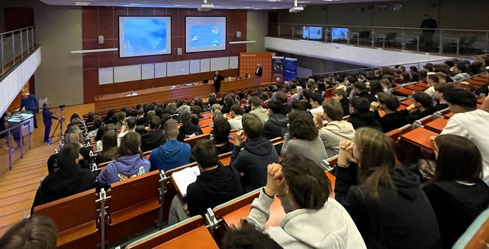 Duża sala wykładowa wypełniona uczniami i uczestnikami wydarzenia. Wszyscy siedzą w rzędach skierowanych w stronę sceny, gdzie prowadzący prezentuje materiał wyświetlany na dwóch dużych ekranach. Widać jasne oświetlenie, drewniane ławki i szeroką przestrzeń audytorium. Uczestnicy uważnie śledzą przebieg wykładu.