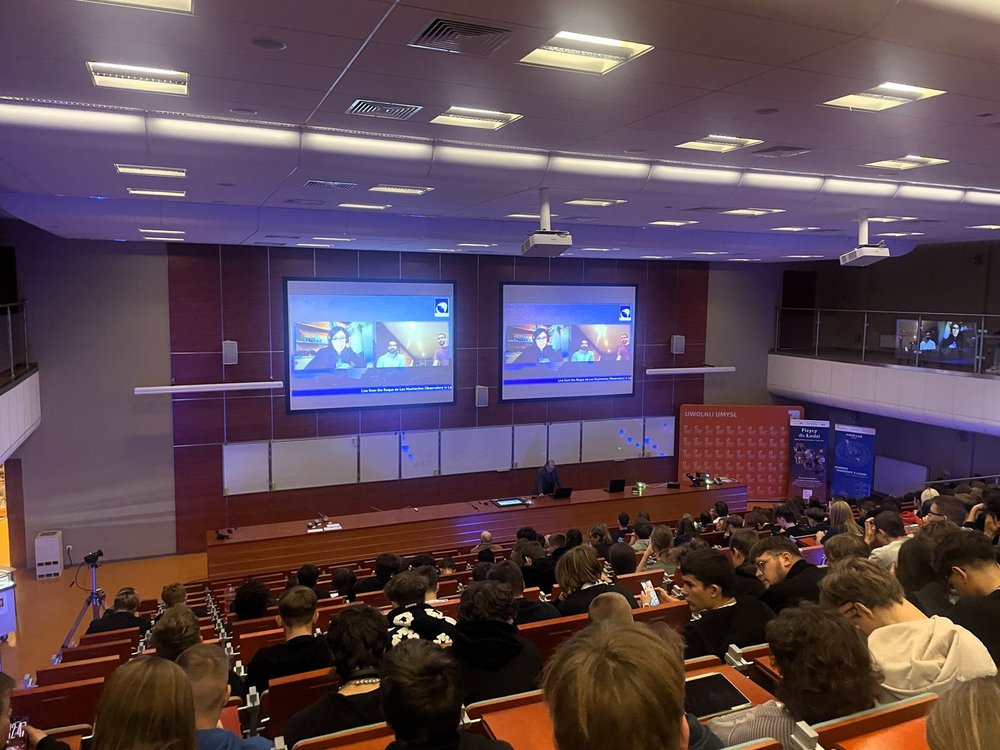 Duża sala wykładowa wypełniona uczniami i uczestnikami wydarzenia. Wszyscy siedzą w rzędach skierowanych w stronę sceny, gdzie prowadzący prezentuje materiał wyświetlany na dwóch dużych ekranach. Widać jasne oświetlenie, drewniane ławki i szeroką przestrzeń audytorium. Uczestnicy uważnie śledzą przebieg wykładu.