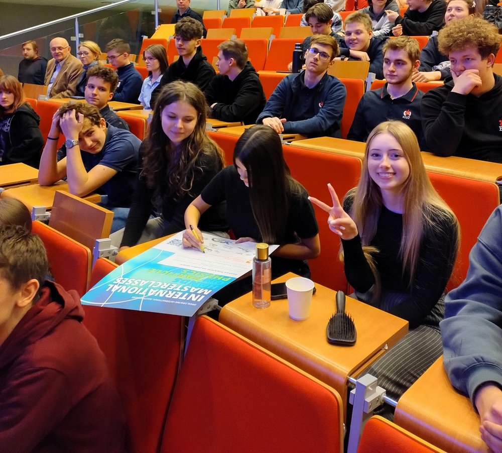 Na zdjęciu widać grupę młodych osób siedzących w rzędach w sali wykładowej z pomarańczowymi fotelami i drewnianymi pulpitami. Na pierwszym planie kilka uczennic i uczniów rozmawia i wypełnia dokument leżący na pulpicie. Jedna osoba pisze na dużej karcie, obok której widać plakat z napisem „International Masterclasses – Physics”. Obok stoi butelka z wodą i papierowy kubek. Po prawej stronie uśmiechnięta dziewczyna patrzy w stronę aparatu i pokazuje gest „V” palcami. W tle siedzi więcej uczestników wydarzenia oraz kilka starszych osób, prawdopodobnie opiekunów lub prowadzących. Atmosfera przypomina spotkanie edukacyjne lub warsztaty na uczelni.