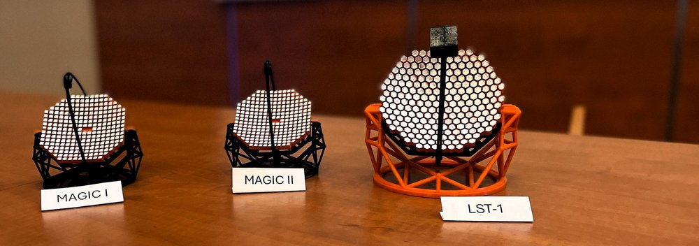 Na drewnianym blacie stoją trzy modele teleskopów Cherenkova. Każdy z nich ma jasną, mozaikową powierzchnię odbijającą oraz konstrukcję przypominającą rusztowanie. Od lewej znajdują się: mały model podpisany ‘MAGIC I’, obok niego podobny model ‘MAGIC II’, a po prawej największy model o strukturze złożonej z sześciokątnych elementów, opisany jako ‘LST-1’. Tło jest rozmyte, co podkreśla detale modeli
