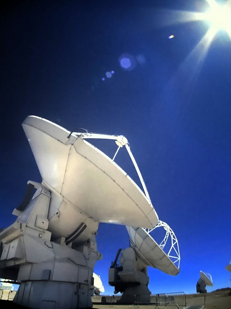 Na zdjęciu widać kilka dużych, białych anten satelitarnych lub radioteleskopów ustawionych na pustynnym, jasnym terenie. Ich talerze skierowane są w niebo. Konstrukcje są masywne i stoją na ciężkich, metalowych podstawach. Niebo jest bardzo czyste, intensywnie granatowe, a w prawym górnym rogu kadru znajduje się oślepiająco jasne Słońce, od którego rozchodzą się promienie światła. W kadrze widoczne są również małe odbłyski – tzw. flary – powstałe od silnego światła słonecznego. Całość sprawia wrażenie wykonanej w wysokogórskim lub pustynnym obserwatorium astronomicznym.