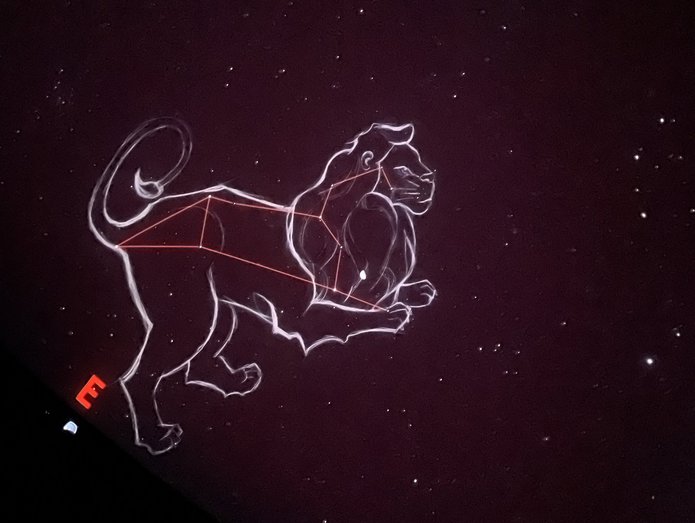 Na ciemnym, gwiaździstym tle widoczna jest ilustracja konstelacji Lwa. Gwiazdy połączone są cienkimi, czerwonymi liniami tworzącymi charakterystyczny kształt, a całość otacza jasny, szkicowy kontur przedstawiający lwa w pozycji stojącej. Po lewej stronie znajduje się czerwona litera „E”, oznaczająca kierunek wschodni. Tło jest jednolicie ciemne, a rozproszone punkty światła przedstawiają gwiazdy.