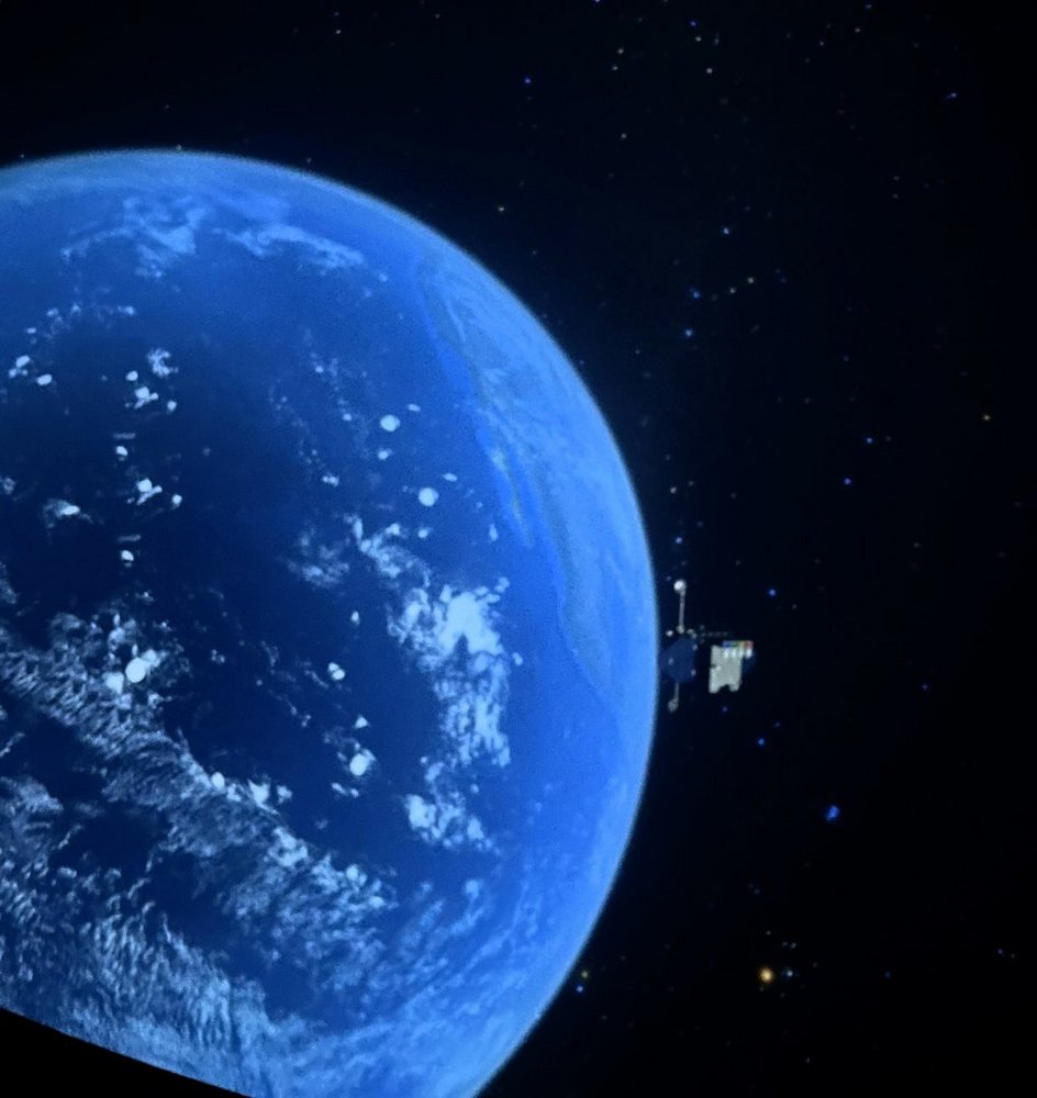 Zdjęcie przedstawia fragment Ziemi widzianej z przestrzeni kosmicznej. Widzimy dużą, zakrzywioną powierzchnię planety, w większości pokrytą oceanem o intensywnie niebieskim kolorze. Na powierzchni widać białe chmury, rozproszone w różnych miejscach nad wodą.  Tło jest czarne, pełne drobnych, jasnych punktów przypominających gwiazdy. Po prawej stronie zdjęcia, na tle gwiazd, znajduje się niewielki obiekt – wygląda jak sztuczny satelita z panelami słonecznymi lub antenami wystającymi z jego boku.  Całość sprawia wrażenie spokojnej sceny obserwowanej z orbity okołoziemskiej.