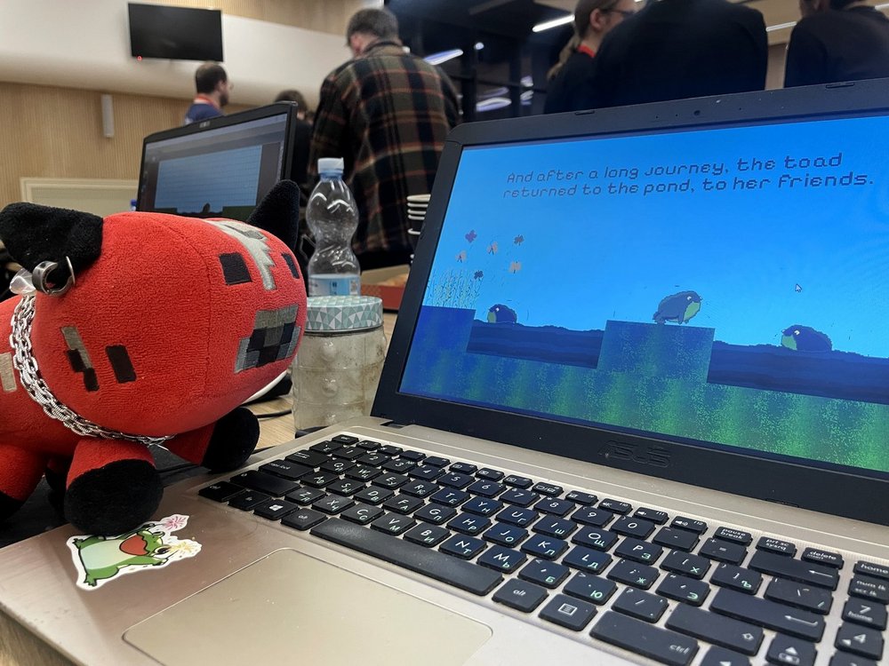 Na zdjęciu widać otwarty laptop stojący na stole podczas wydarzenia. Na ekranie wyświetlana jest gra komputerowa w stylu pikselowym z żabą na tle nieba i wody oraz tekstem końcowym gry. Obok laptopa leży czerwono-czarna pluszowa maskotka w kształcie zwierzątka z kolczykiem i łańcuszkiem. Na stole znajdują się także butelka wody i słoik. W tle widać grupę osób stojących w sali, co sugeruje, że zdjęcie zostało wykonane podczas maratonu tworzenia gier.