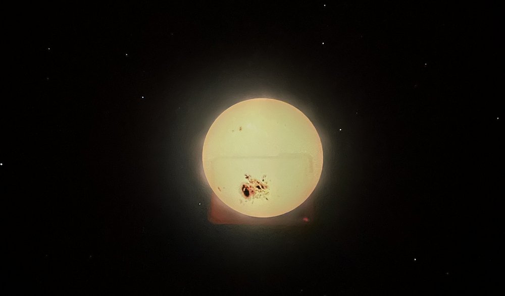 Na środku obrazu znajduje się duża, jasnożółta kula przedstawiająca Słońce. Jego powierzchnia jest nierówna i dynamiczna – widoczne są jaśniejsze i ciemniejsze obszary, przypominające gorące, wijące się struktury plazmy. Tło zdjęcia jest czarne i wypełnione licznymi gwiazdami w różnych kolorach: białymi, niebieskimi, pomarańczowymi i czerwonymi. 