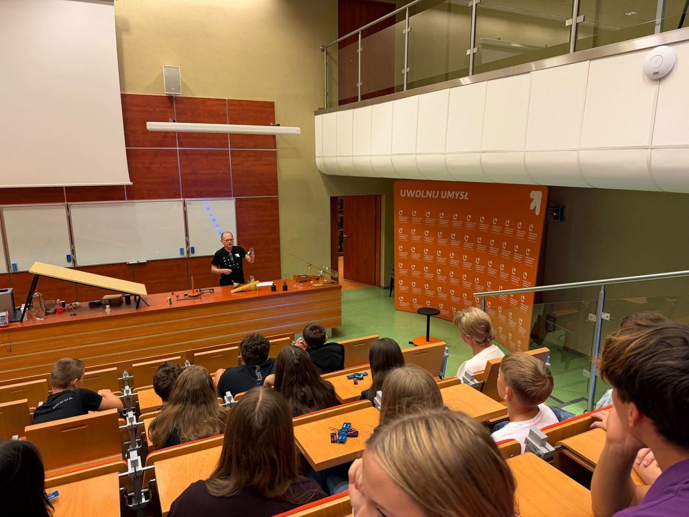 Zdjęcie przedstawia salę wykładową podczas pokazu doświadczeń fizycznych. Przy stole demonstracyjnym stoi prowadzący prezentujący eksperyment, a przed nim siedzi grupa uczniów obserwujących pokaz. Na stołach i stanowisku laboratoryjnym widoczne są różne przyrządy i zestawy do doświadczeń. W tle znajduje się tablica, ekran projekcyjny oraz charakterystyczna pomarańczowa ścianka z identyfikacją wizualną Wydziału.