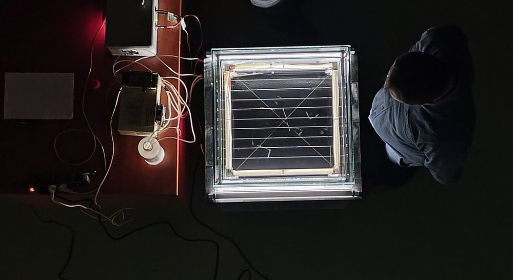 Zdjęcie wykonane z góry przedstawia stanowisko laboratoryjne w ciemnym pomieszczeniu. W centrum znajduje się podświetlone, kwadratowe urządzenie z przezroczystą obudową i jasną ramką świetlną. W jego wnętrzu widać cienkie linie i elementy przypominające układ pomiarowy lub eksperymentalny. Po lewej stronie znajduje się stół z różnymi urządzeniami elektronicznymi, przewodami oraz małą lampką. Po prawej stronie stoi osoba pochylona nad stanowiskiem i obserwująca eksperyment. Całość wygląda jak demonstracja lub badanie prowadzone w laboratorium fizycznym.