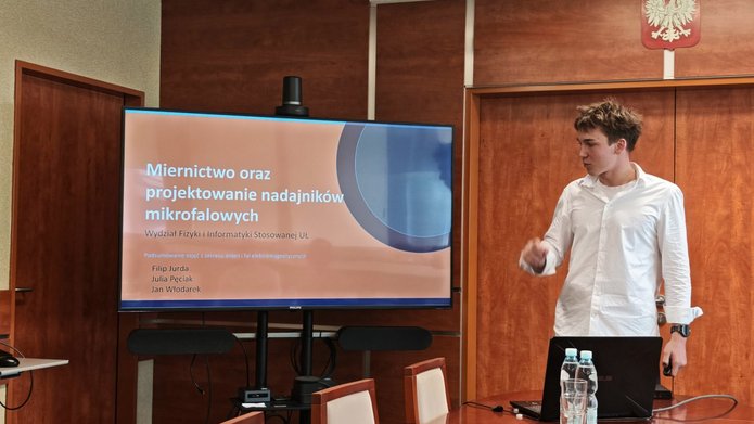 W sali konferencyjnej młody mężczyzna prowadzi prezentację przy dużym ekranie multimedialnym. Ubrany jest w białą koszulę i stoi po prawej stronie ekranu, zwrócony w jego kierunku. Na ekranie wyświetlony jest slajd tytułowy prezentacji zatytułowanej „Miernictwo oraz projektowanie nadajników mikrofalowych”. Przed prelegentem znajduje się stół konferencyjny z otwartym laptopem, butelkami wody, szklankami i talerzykiem z poczęstunkiem. W tle widoczne są drewniane ściany oraz godło Polski umieszczone wysoko nad drzwiami. Zdjęcie przedstawia formalne wystąpienie podczas wydarzenia edukacyjnego lub naukowego.