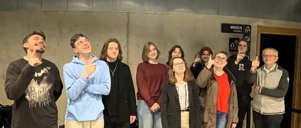 Zdjęcie przedstawia grupę dziesięciu osób stojących w dwóch rzędach na tle szarej, betonowej ściany. Większość z nich uśmiecha się i wskazuje palcem w górę, jakby patrzyli na coś nad sobą. W pierwszym rzędzie stoją osoby w swobodnych, codziennych ubraniach: po lewej osoba w ciemnej bluzie, obok osoba w jasnej, niebieskiej bluzie, dalej kilka osób w stonowanych kolorach — czerni, bordzie i beżu. W tylnym rzędzie widać trzy osoby również wykonujące gest wskazywania w górę. Po prawej stronie stoi starszy mężczyzna w szarej bluzie, także wskazujący palcem do góry. W tle znajduje się wejście oznaczone czarno-białą tabliczką.