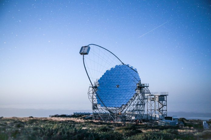 Pojedynczy teleskop astronomiczny z dużym, segmentowym zwierciadłem i stalową konstrukcją wsporczą na górskim szczycie. Nad teleskopem rozciąga się błękitne, rozgwieżdżone niebo, a w tle widoczna jest warstwa chmur.