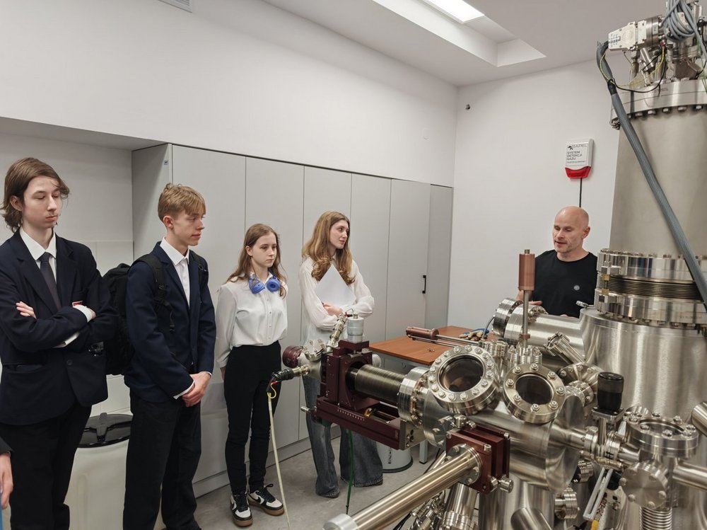 W nowoczesnym laboratorium grupa czworga młodych ludzi słucha wyjaśnień prowadzącego. Po prawej stronie stoi mężczyzna, który omawia działanie dużej, metalowej aparatury badawczej. Uczestnicy stoją obok siebie i z uwagą obserwują urządzenie. W pomieszczeniu dominują jasne ściany oraz specjalistyczna aparatura wykonana ze stali nierdzewnej. Zdjęcie przedstawia wizytę edukacyjną lub zajęcia laboratoryjne związane z naukami ścisłymi.