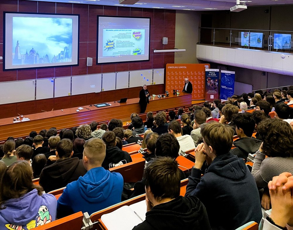 Duża sala wykładowa wypełniona uczniami i uczestnikami wydarzenia. Wszyscy siedzą w rzędach skierowanych w stronę sceny, gdzie prowadzący prezentuje materiał wyświetlany na dwóch dużych ekranach. Widać jasne oświetlenie, drewniane ławki i szeroką przestrzeń audytorium. Uczestnicy uważnie śledzą przebieg wykładu.