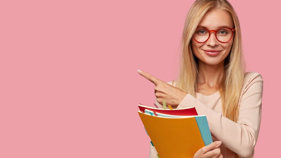 Studio shot of good looking Caucasian teacher attracts your attention to copy space, holds books, wears round spectacles, explains material to pupils, points with index finger on pink background Zdjęcie-uśmiechnięta studentka trzyma trzy kolorowe zeszyty