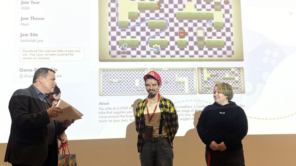 Na zdjęciu widać trzy osoby stojące przed dużym ekranem z wyświetloną prezentacją gry komputerowej zatytułowanej „Shortness of Breath”. Po lewej stronie stoi Tomasz Gwizdałła i  ogłasza wyniki oraz wręcza nagrody. Pośrodku stoi młody mężczyzna w czerwonej czapce i koszuli w kratę, z identyfikatorem na szyi. Po prawej stronie stoi młoda kobieta ubrana na czarno.