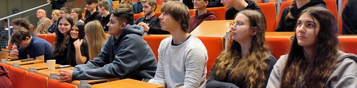 Na zdjęciu widać grupę młodych osób siedzących w rzędach w dużej sali wykładowej. Młodzież zajmuje miejsca przy drewnianych pulpitach i siedzą na pomarańczowych fotelach ustawionych schodkowo. Większość patrzy w kierunku prowadzącego znajdującego się poza kadrem, słuchając wykładu lub prezentacji. Na pierwszym planie widać kilka osób siedzących obok siebie – część z nich opiera ręce na blatach, jedna osoba trzyma kubek. W tle widoczni są kolejni uczestnicy spotkania oraz starszy mężczyzna siedzący wyżej w rzędzie. Atmosfera przypomina zajęcia lub warsztaty akademickie.