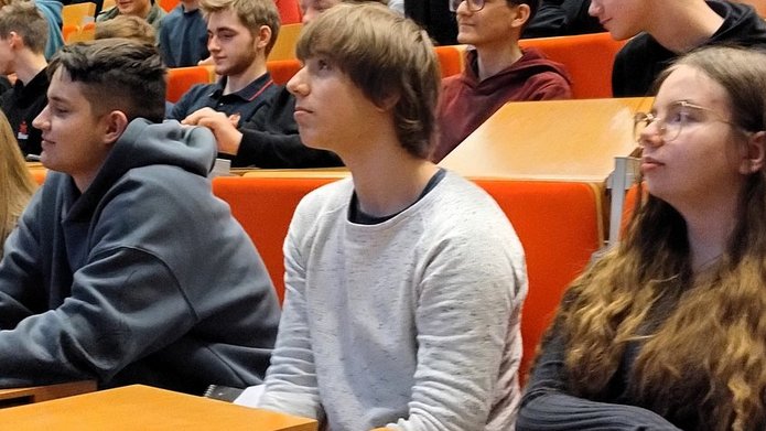 Na zdjęciu widać grupę młodych osób siedzących w rzędach w dużej sali wykładowej. Młodzież zajmuje miejsca przy drewnianych pulpitach i siedzą na pomarańczowych fotelach ustawionych schodkowo. Większość patrzy w kierunku prowadzącego znajdującego się poza kadrem, słuchając wykładu lub prezentacji. Na pierwszym planie widać kilka osób siedzących obok siebie – część z nich opiera ręce na blatach, jedna osoba trzyma kubek. W tle widoczni są kolejni uczestnicy spotkania oraz starszy mężczyzna siedzący wyżej w rzędzie. Atmosfera przypomina zajęcia lub warsztaty akademickie.