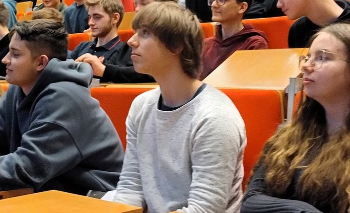 Na zdjęciu widać grupę młodych osób siedzących w rzędach w dużej sali wykładowej. Młodzież zajmuje miejsca przy drewnianych pulpitach i siedzą na pomarańczowych fotelach ustawionych schodkowo. Większość patrzy w kierunku prowadzącego znajdującego się poza kadrem, słuchając wykładu lub prezentacji. Na pierwszym planie widać kilka osób siedzących obok siebie – część z nich opiera ręce na blatach, jedna osoba trzyma kubek. W tle widoczni są kolejni uczestnicy spotkania oraz starszy mężczyzna siedzący wyżej w rzędzie. Atmosfera przypomina zajęcia lub warsztaty akademickie.