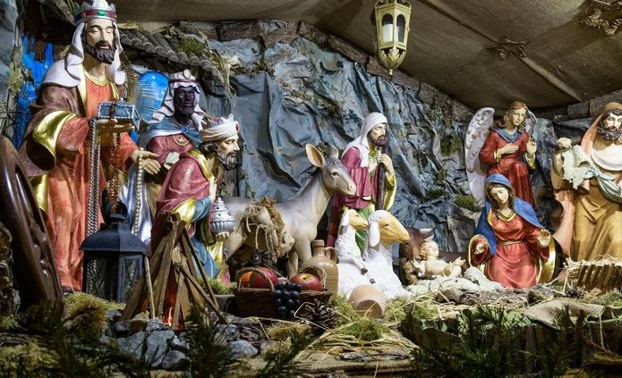 Na obrazie widoczna jest szopka bożonarodzeniowa z wieloma figurami. Scena przedstawia narodzenie Jezusa.  W środku leży małe dziecko – Jezus, ułożone na jasnym sianie w drewnianym żłóbku. Dziecko ma jasną skórę, jest nagie, owinięte delikatną tkaniną. Ręce ma lekko uniesione.  Po prawej stronie dziecka klęczy Maryja. Ma spokojną twarz i spuszczony wzrok. Jest ubrana w czerwoną suknię i niebieski płaszcz. Ręce ma złożone w geście modlitwy.  Obok Maryi stoi święty Józef. Jest ubrany w jasną szatę, ma brodę i trzyma baranka na rękach. Jego twarz jest łagodna.  Po lewej stronie znajdują się Trzej Królowie. To trzej mężczyźni w bogatych, kolorowych strojach. Jeden z nich klęczy i trzyma naczynie z darem. Pozostali stoją, trzymając prezenty dla dziecka. Jeden z Królów ma ciemną skórę, pozostali jaśniejszą.  Za nimi widać zwierzęta: osła, owce i baranki. Zwierzęta stoją spokojnie, zwrócone w stronę dziecka.  W tle znajduje się skalna grota i fragmenty drewnianej konstrukcji. Nad postaciami wiszą lampy i gwiazdy, które oświetlają scenę ciepłym światłem.  Całość ma uroczysty i spokojny charakter, a kolory są ciepłe: brązy, złoto, czerwień i niebieski. Scena symbolizuje narodziny Jezusa i moment oddania czci przez ludzi i zwierzęta.