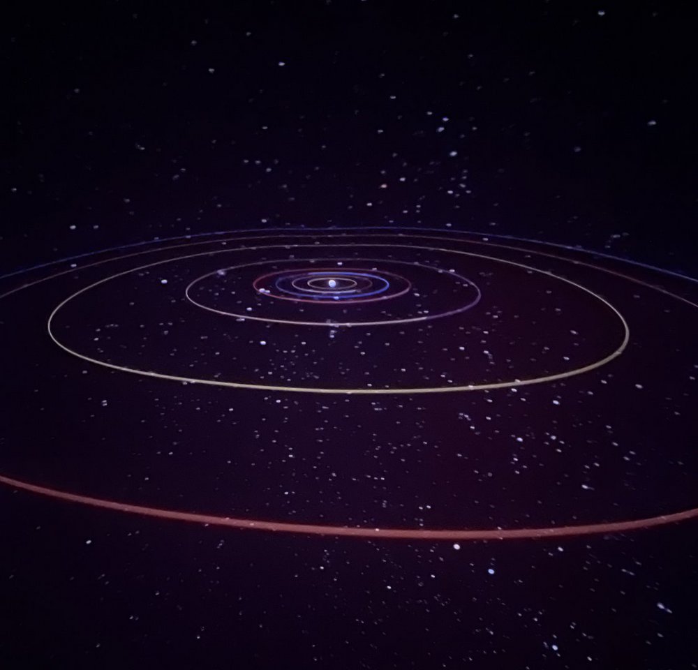 Zdjęcie przedstawia wizualizację Układu Słonecznego na tle ciemnego, gwiaździstego nieba. W centrum widoczny jest jasny punkt symbolizujący Słońce, a wokół niego rozchodzą się koncentryczne, kolorowe linie przedstawiające orbity planet. Linie mają różne barwy – od jasnych, przez niebieskie i fioletowe, po czerwone – tworząc wielowarstwowy, okrągły układ pośród rozproszonych białych punktów imitujących gwiazdy.