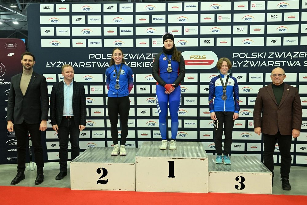 Na zdjęciu widoczna jest ceremonia dekoracji podczas zawodów łyżwiarstwa szybkiego. Na podium stoją trzy młode zawodniczki w strojach sportowych. Zawodniczka na najwyższym stopniu podium (miejsce 1) ubrana jest w niebieski strój startowy i trzyma złoty medal. Po jej lewej stronie stoi zawodniczka na miejscu 2 – ma na sobie niebieską bluzę klubową i czarne legginsy, a na szyi srebrny medal. Po prawej stronie podium stoi zawodniczka z miejsca 3, również w stroju sportowym, z brązowym medalem. Za podium znajduje się ścianka z logotypami sponsorów i organizatorów zawodów. Po obu stronach zawodniczek stoją czterej mężczyźni w eleganckich ubraniach, prawdopodobnie przedstawiciele organizatorów wręczający nagrody. Scena odbywa się na czerwonym dywanie w hali sportowej.